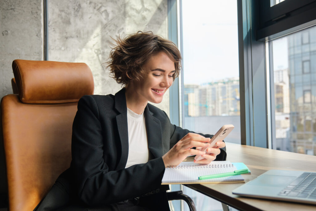 Portrait of corporate woman in suit sits in office messaging someone replying to friend on smartphone working with laptop having lunch break - Bidaya Global